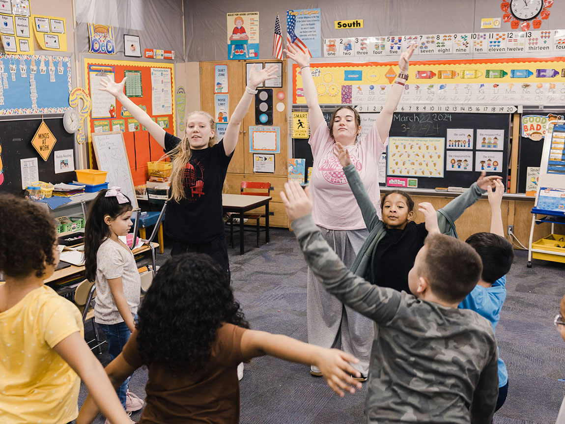 Two college students lead elementary school students in a classroom in a warmup before dance class